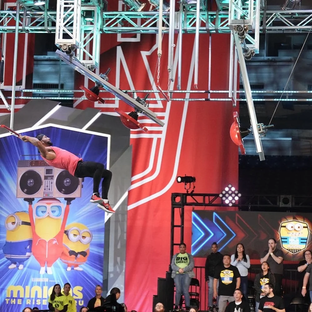 Emmanuel Aouad on the American Ninja Warrior course, mid-obstacle under stage lighting.